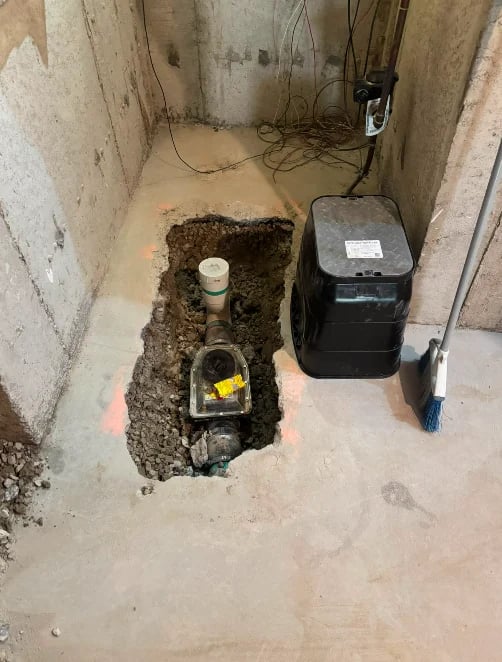 Underground sump pit with submersible pump in concrete basement, black control box mounted on right wall, electrical wiring overhead