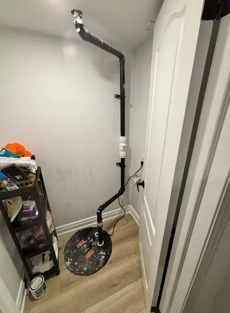 Black drain pipe with elbow joints extending up a white wall from a circular base on wooden floor in a corner closet