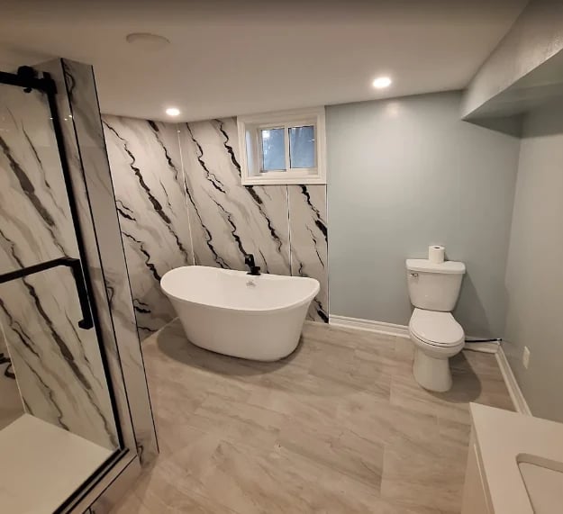 Modern bathroom with white freestanding tub, glass shower enclosure, toilet, and marble-patterned tile accent wall