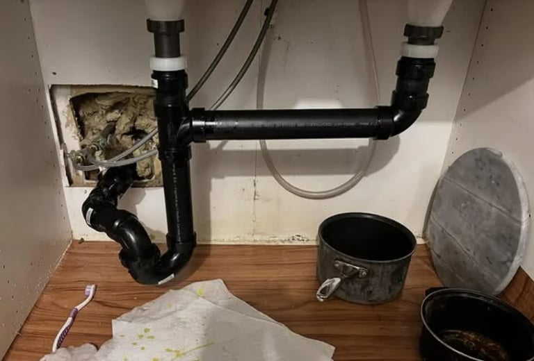 Under-sink plumbing with black P-trap pipes, white electrical outlet box, metal containers, and white papers on wooden floor